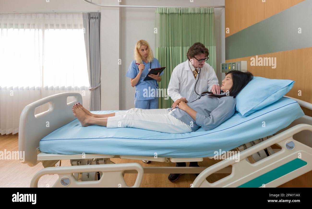 Caucasian doctor holding stethoscope checking heartbeat, examining illness of asian woman patient on the bed in hospital. Young nurse stand next to th Stock Photo