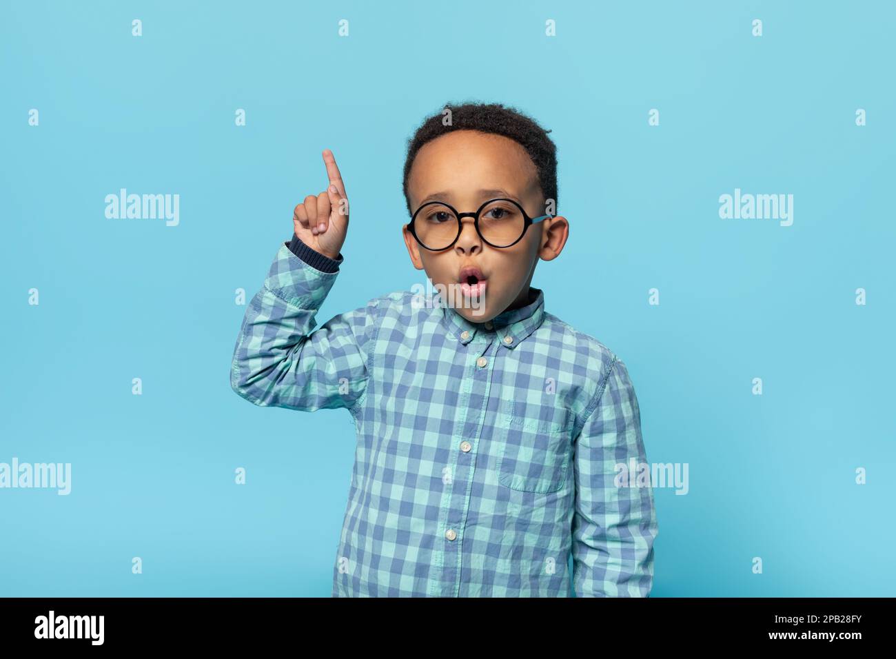 Great idea. Emotional black boy in eyeglasses pointing finger up and looking at camera, posing over blue background Stock Photo