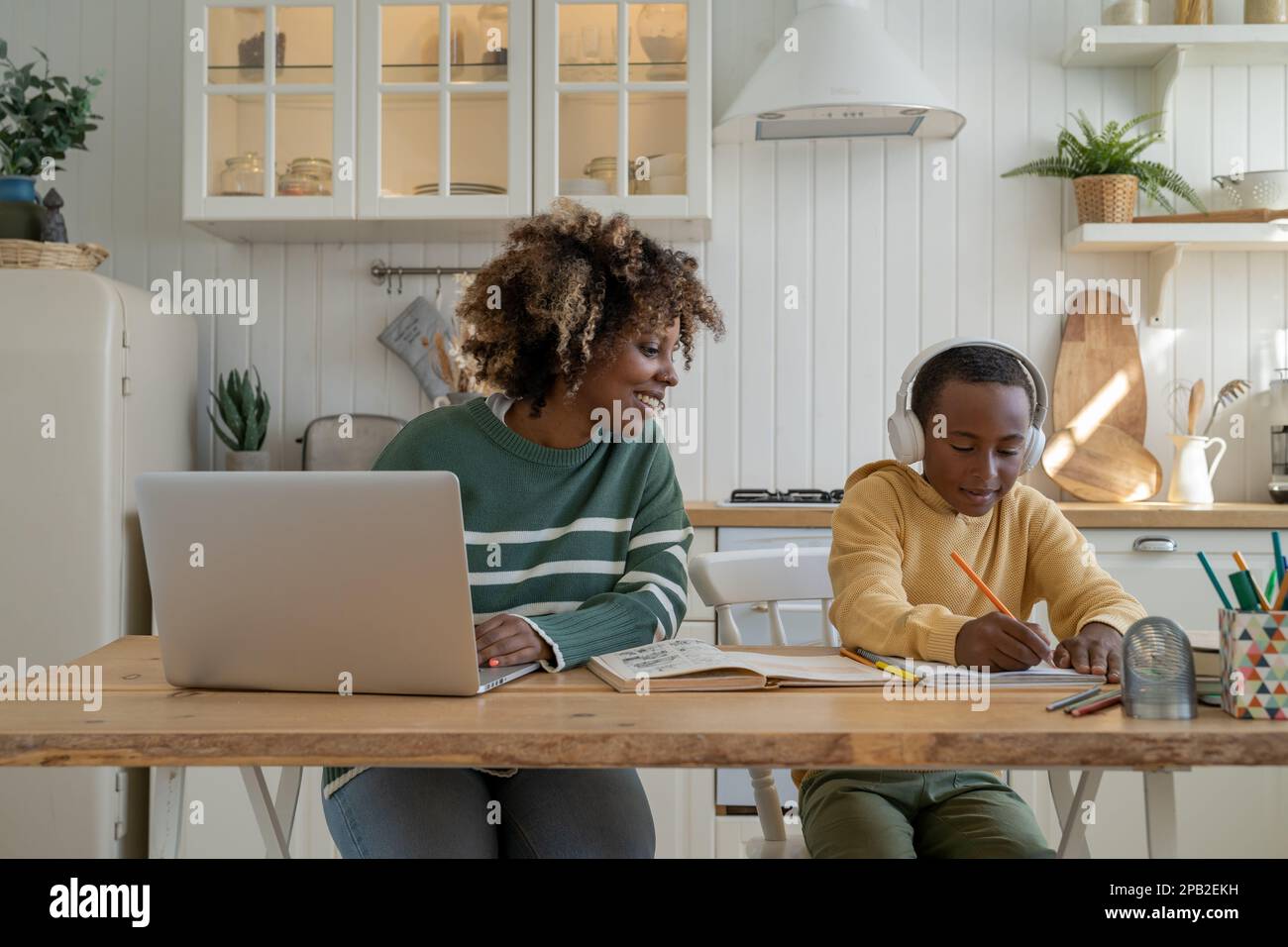 Smiling black mother helping small pensive son with school tasks studying together at kitchen table  Stock Photo