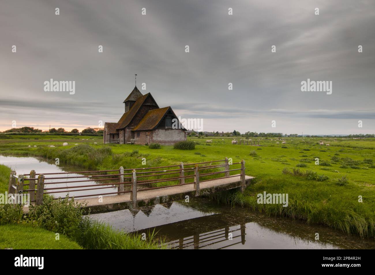 View of sheep on grazing marsh with footbridge across flooded ditch and church in evening, St. Thomas a Becket Church, Fairfield, Walland Marsh, Romne Stock Photo