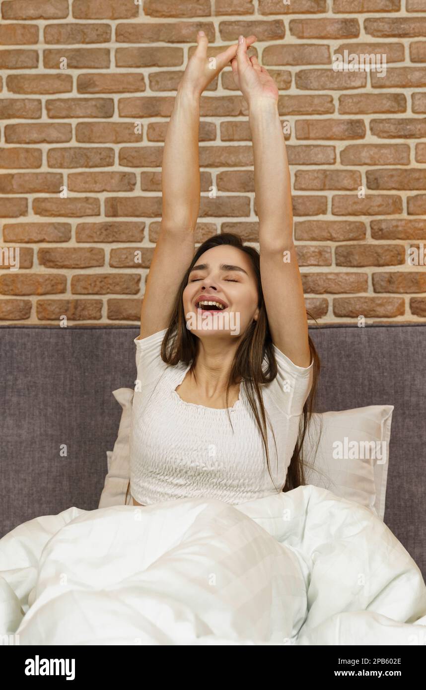 Beautiful woman sits on the bed, stretches her arms up after sleeping ...