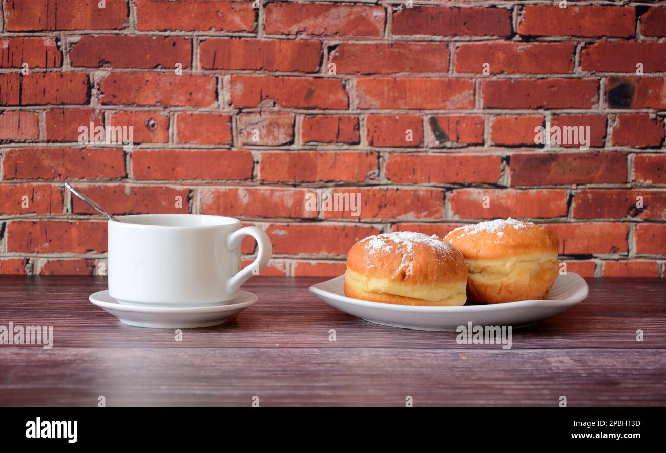 A cup of hot black coffee and a plate with two fresh donuts in powdered sugar on a wooden table against a red brick wall. Close-up. Stock Photo