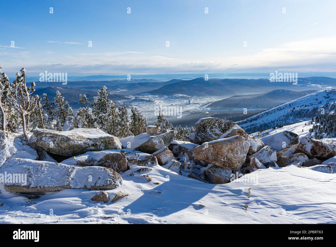Stones in hoarfrost and dwarf conifers in the snow, on top of a high mountain. View of the hills and the village Stock Photo
