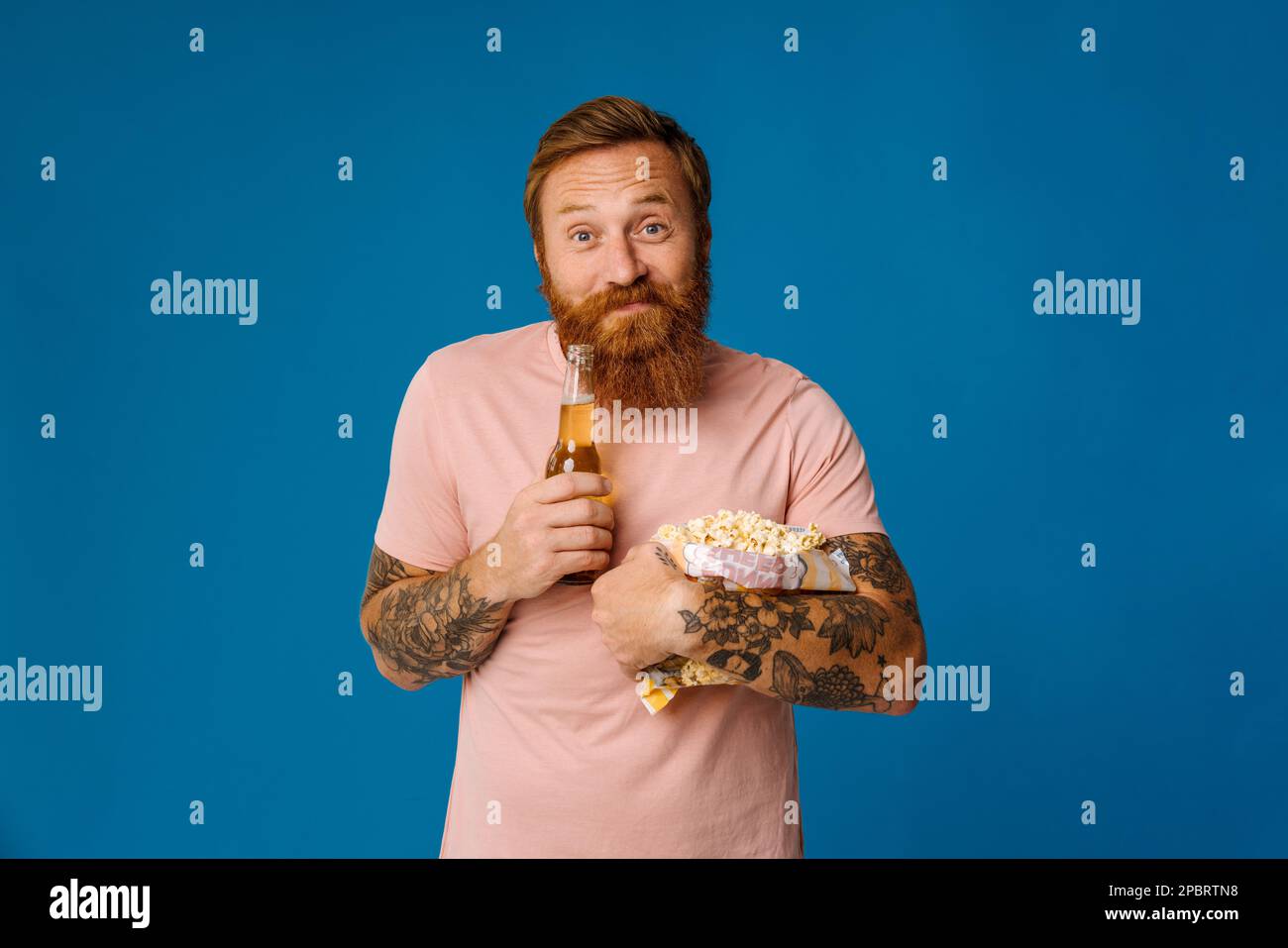 Pleased ginger bearded man holding beer and popcorn while standing isolated over blue studio background Stock Photo