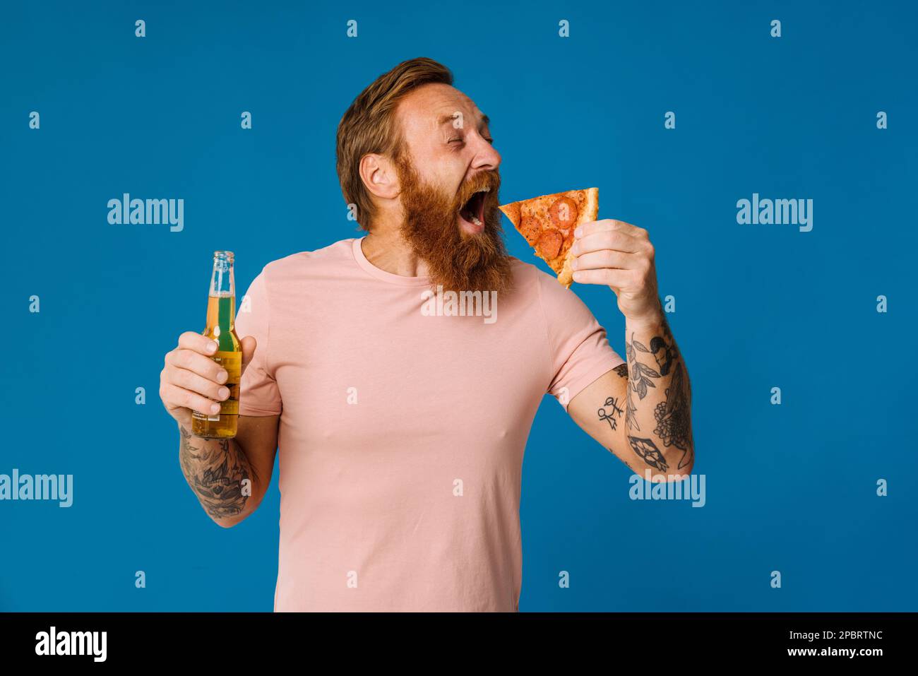Hungry bearded man eating pizza and holding beer isolated over blue background Stock Photo