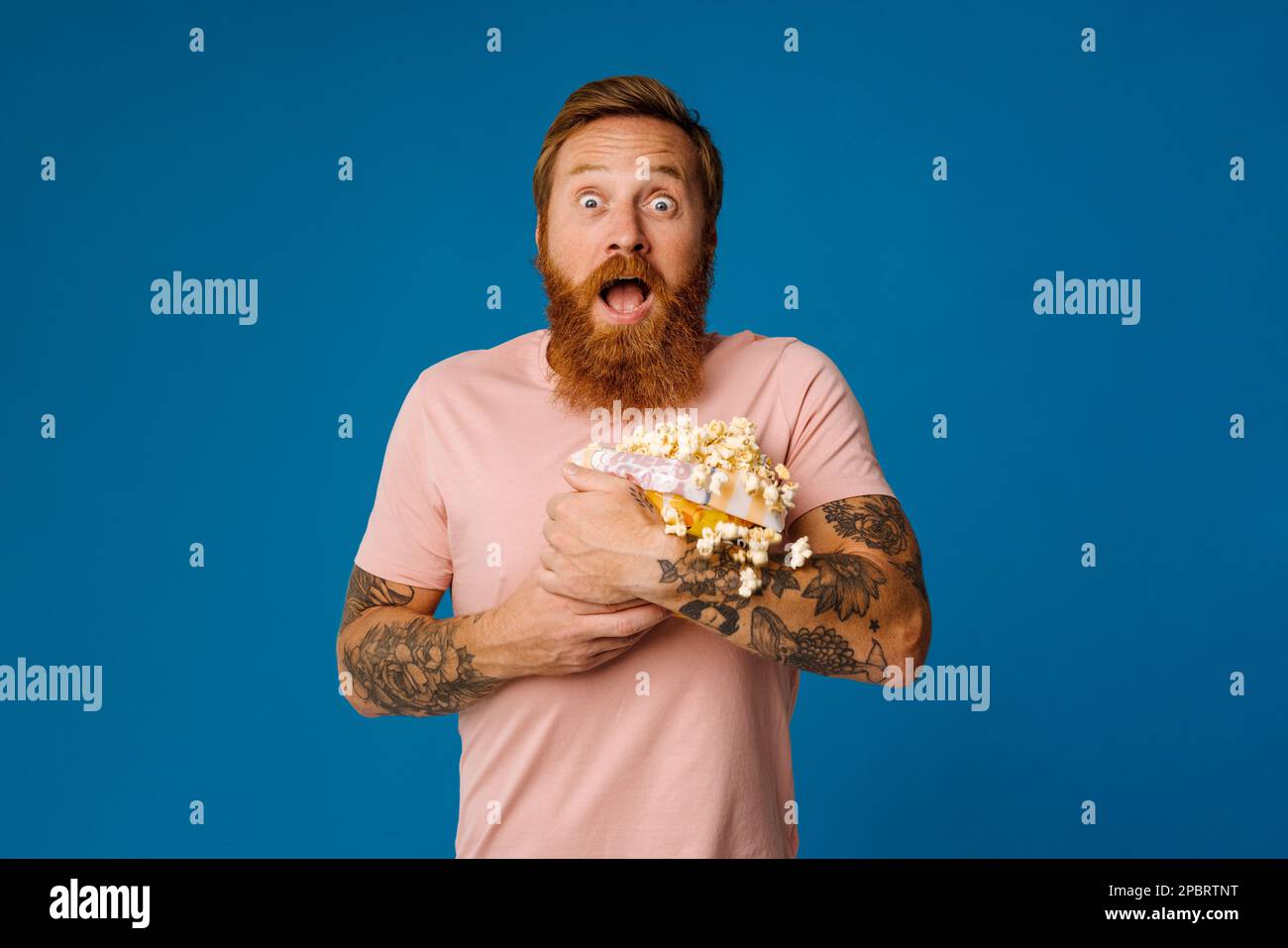 Shocked bearded man holding bucket with flying popcorn isolated over blue studio background Stock Photo