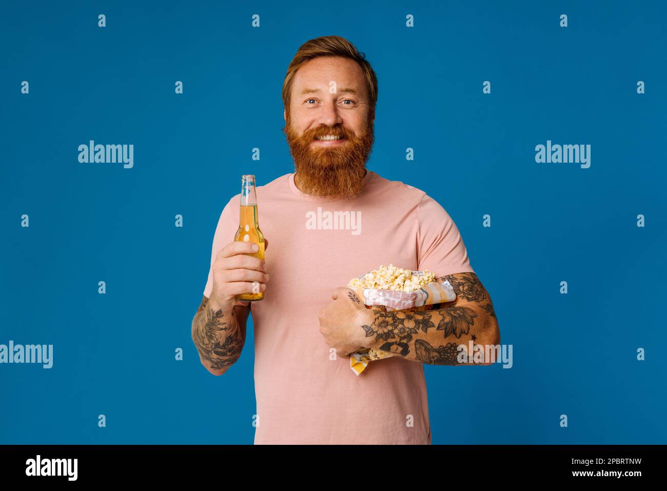 Portrait of excited ginger bearded man holding beer and popcorn while standing isolated over blue studio background Stock Photo