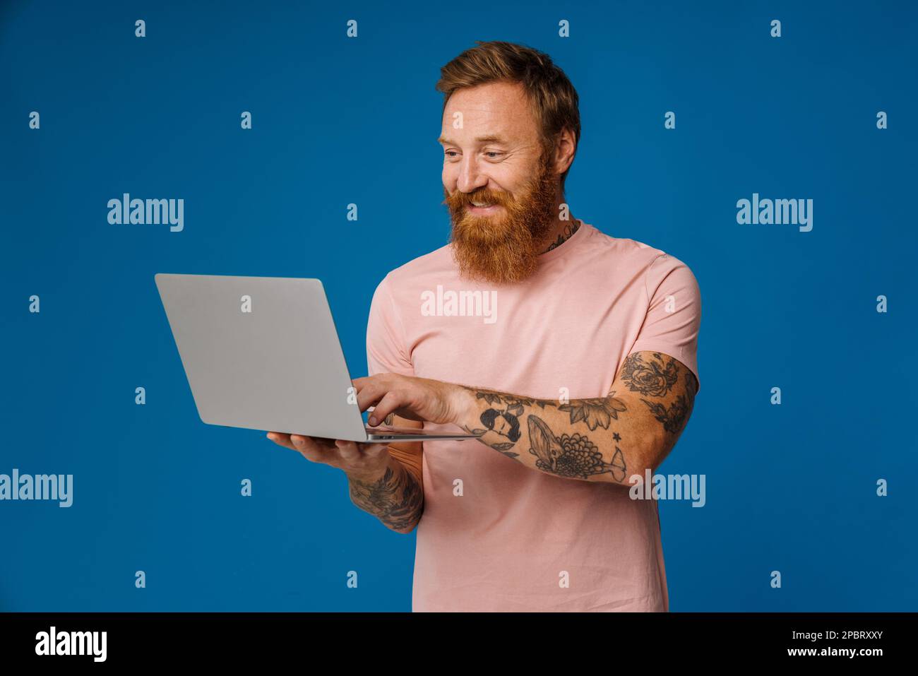 Bearded happy man holding laptop while standing isolated over blue background Stock Photo