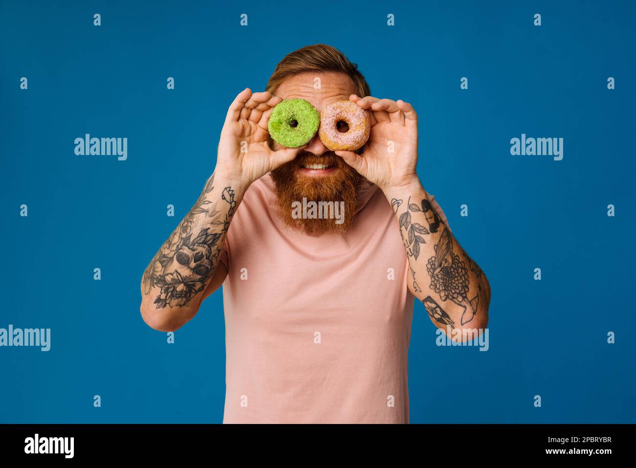 Bearded happy man holding donuts while standing isolated over blue background Stock Photo