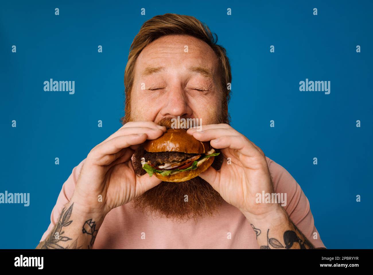 Bearded hungry man eating burger while standing isolated over blue background Stock Photo