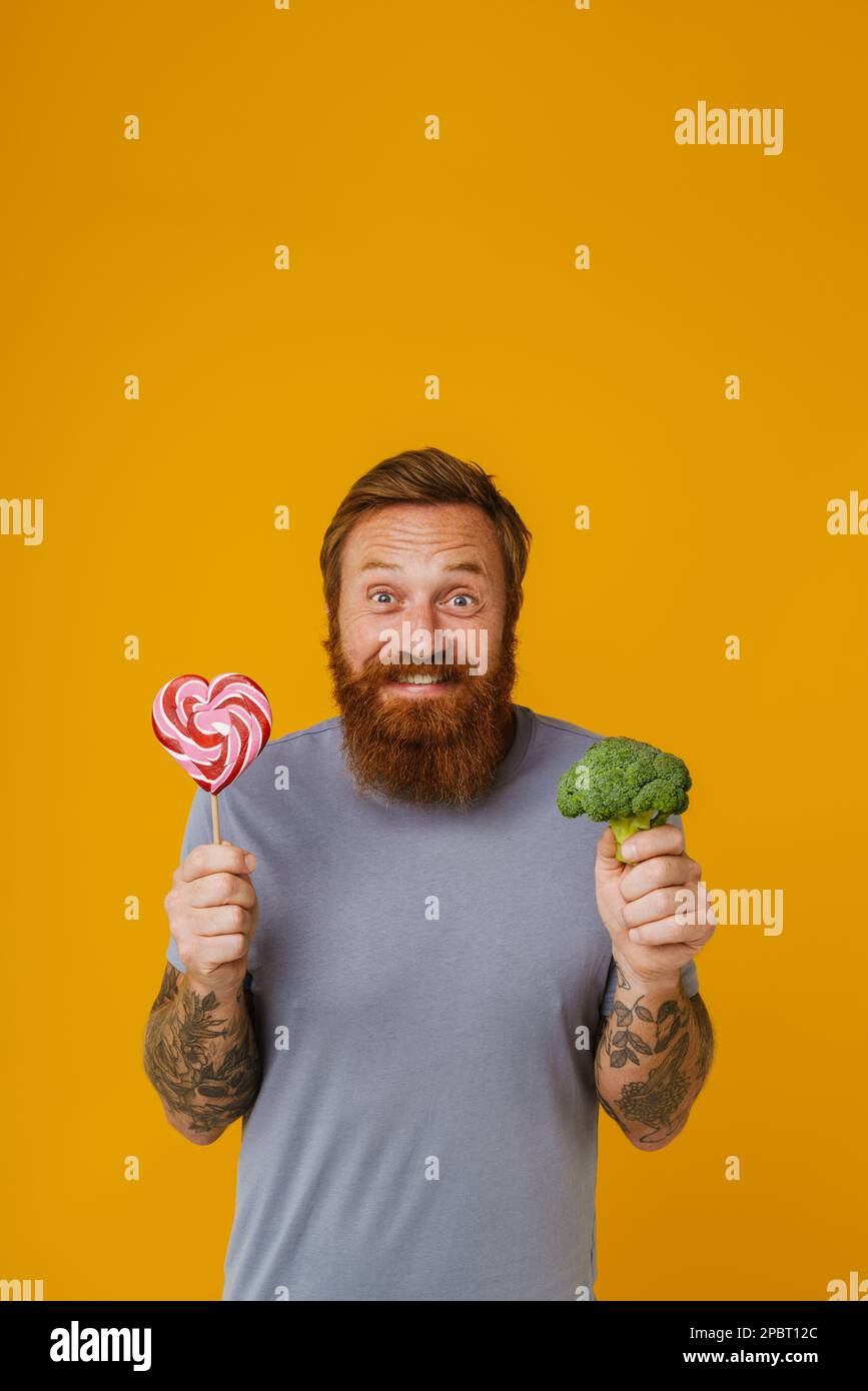 Bearded man holding lollipop and broccoli while standing isolated over yellow background Stock Photo