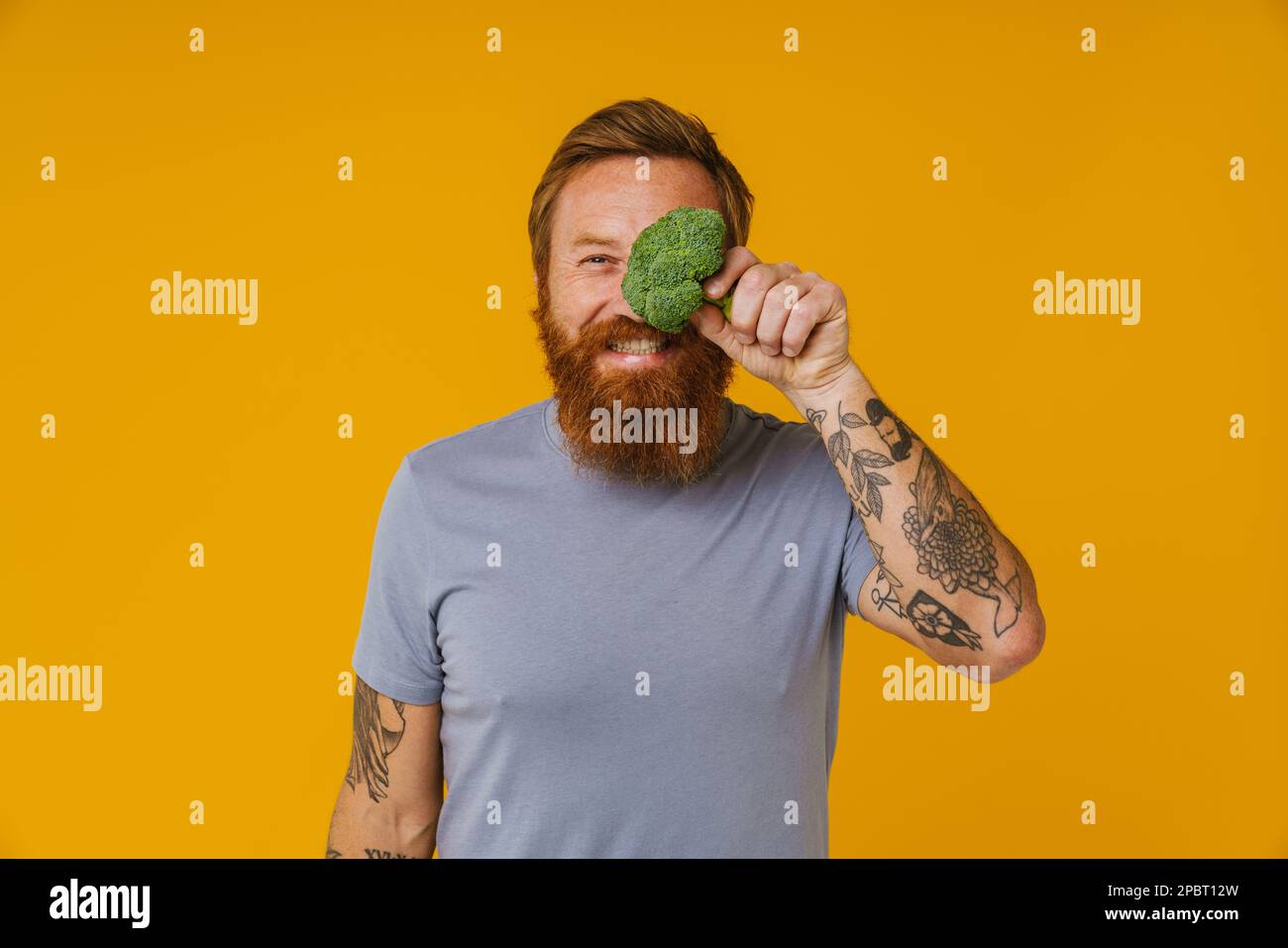 Bearded happy man holding broccoli while standing isolated over yellow background Stock Photo