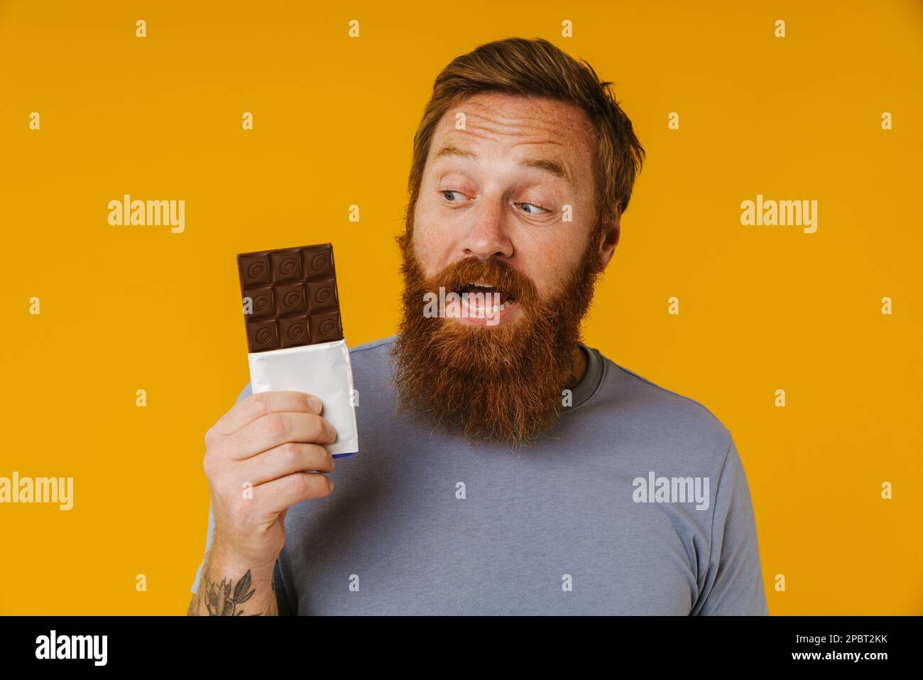 Bearded happy man holding chocolate bar while standing isolated over yellow background Stock Photo