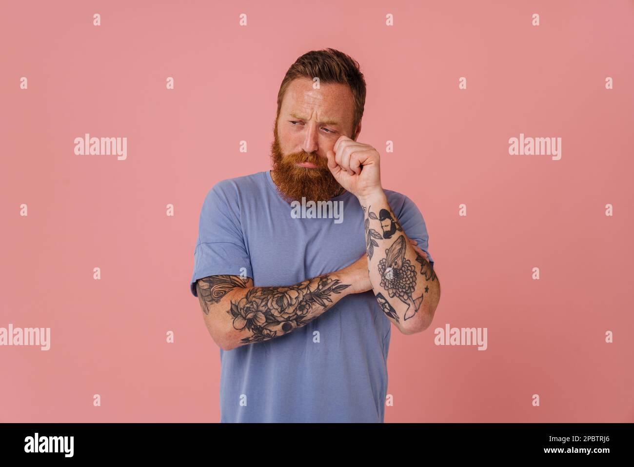 Ginger sad man with beard crying while wiping his tears isolated over pink background Stock Photo