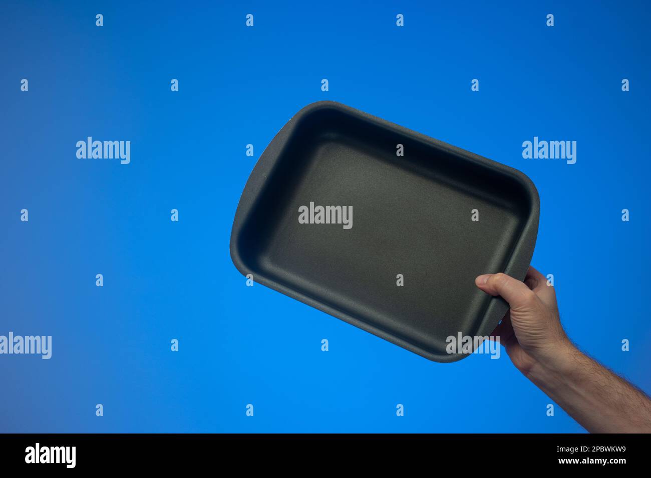 Black nonstick empty rectangular oven tray held by Caucasian male hand. Close up studio shot, isolated on blue background. Stock Photo