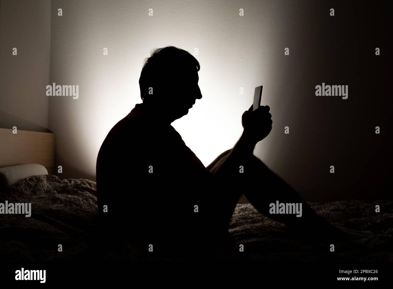 Male silhouette depicting loneliness or depression sitting in his apartment bed, looking at social media on his phone. Stock Photo