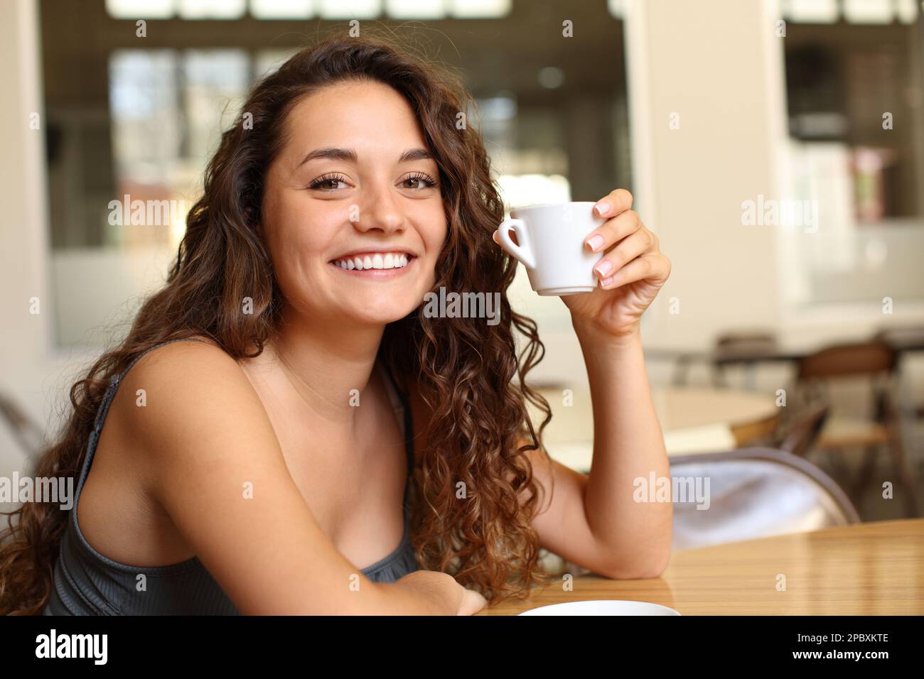 Happy woman holding coffee mug looking at you sitting in a bar and smiling Stock Photo