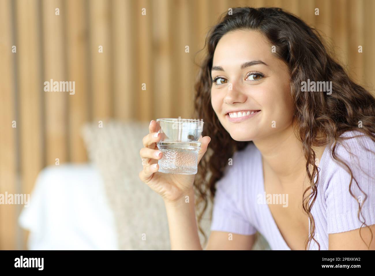 Happy woman holding water glass looks at you sitting on a bed Stock Photo
