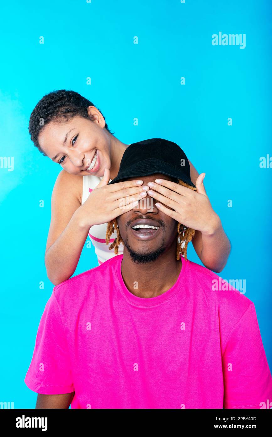 Positive enamored young mixed race lady with dark short hair smiling and looking at camera while covering eyes of cheerful black boyfriend against blu Stock Photo