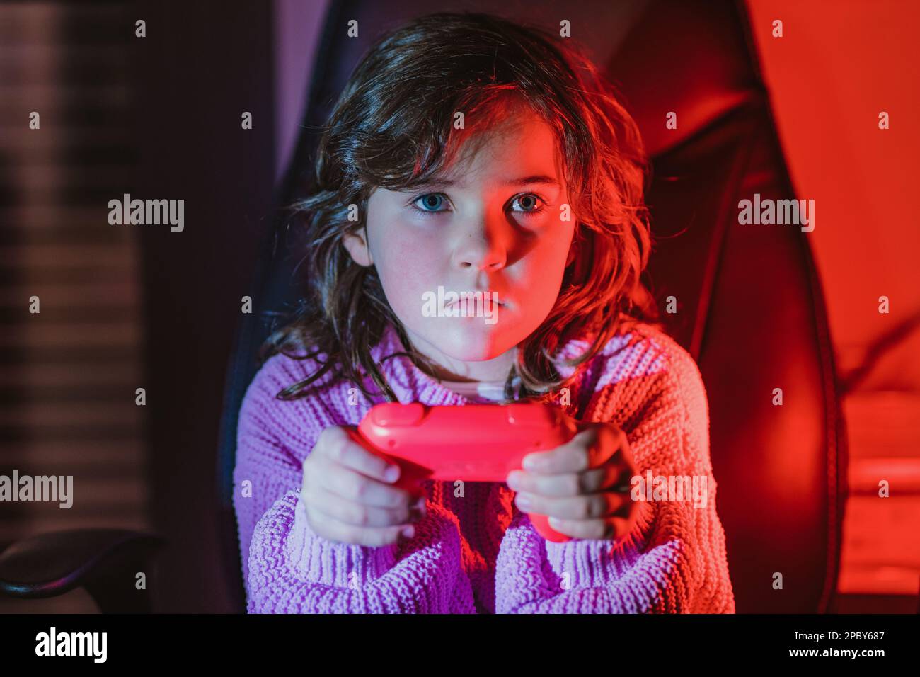 Cute attentive little girl with long brown hair in warm sweater using gamepad while playing videogame sitting on black leather chair in dark room with Stock Photo