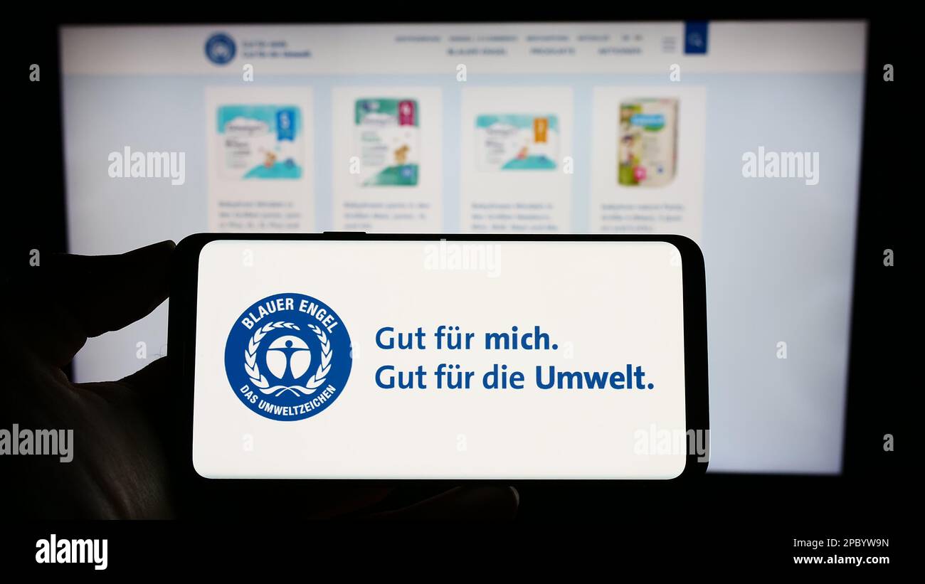 Person holding cellphone with logo of German environmental label Blauer Engel (Blue Angel) on screen in front of webpage. Focus on phone display. Stock Photo