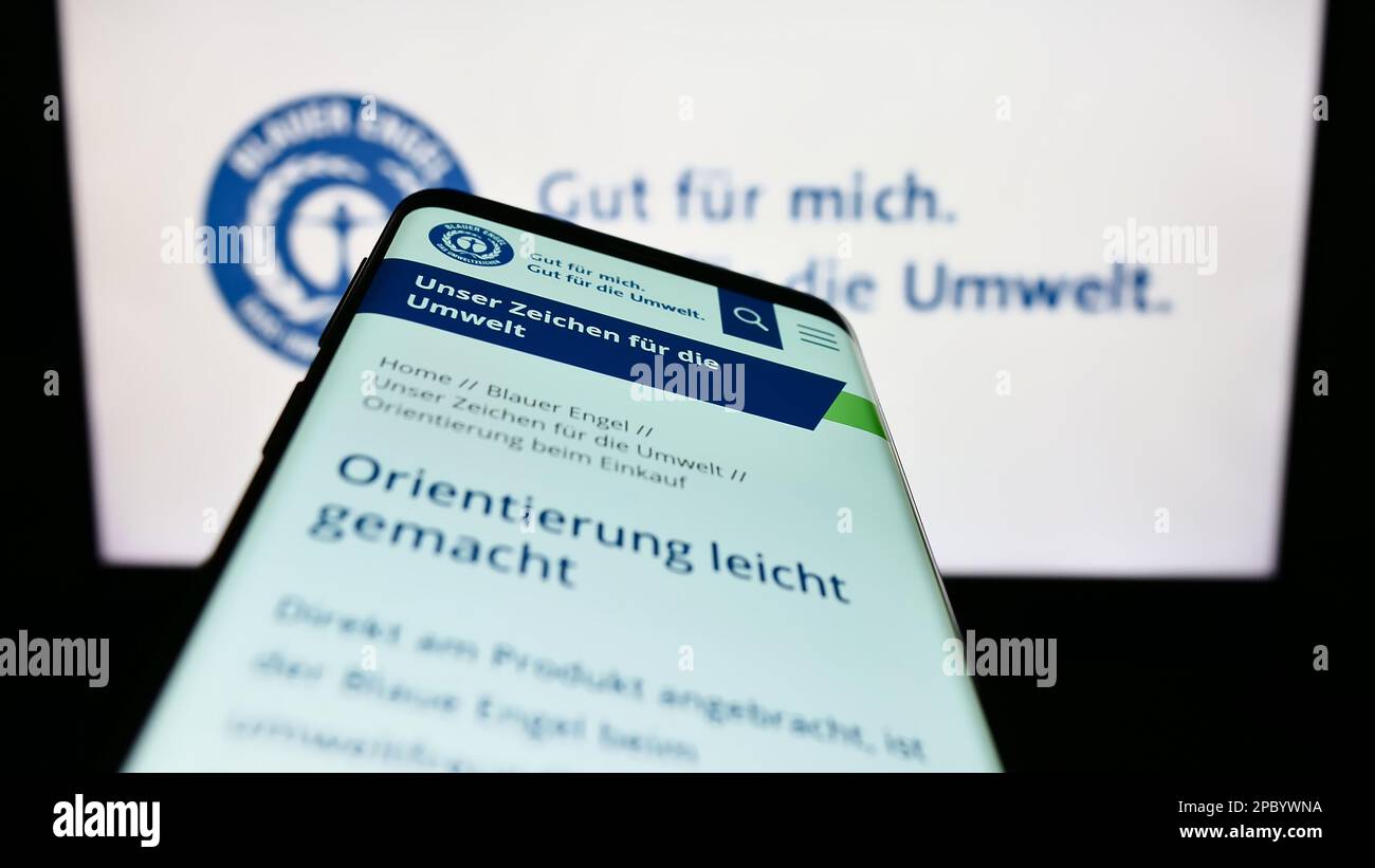 Mobile phone with webpage of German environmental label Blauer Engel (Blue Angel) on screen in front of logo. Focus on top-left of phone display. Stock Photo