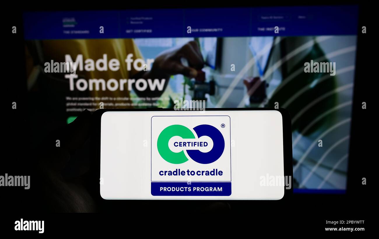 Person holding smartphone with logo of environmental label Cradle to Cradle Certified on screen in front of website. Focus on phone display. Stock Photo