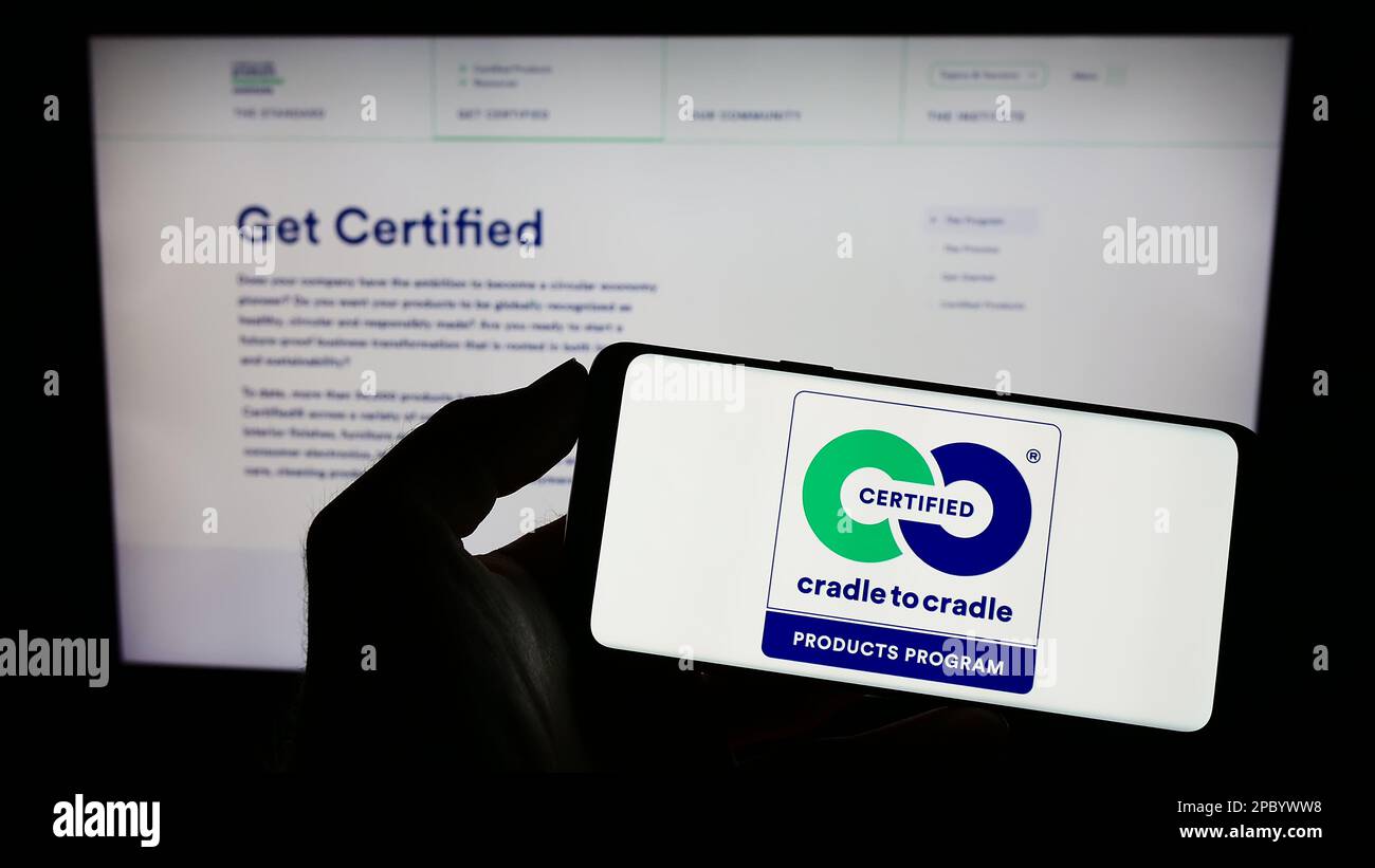 Person holding mobile phone with logo of environmental label Cradle to Cradle Certified on screen in front of web page. Focus on phone display. Stock Photo