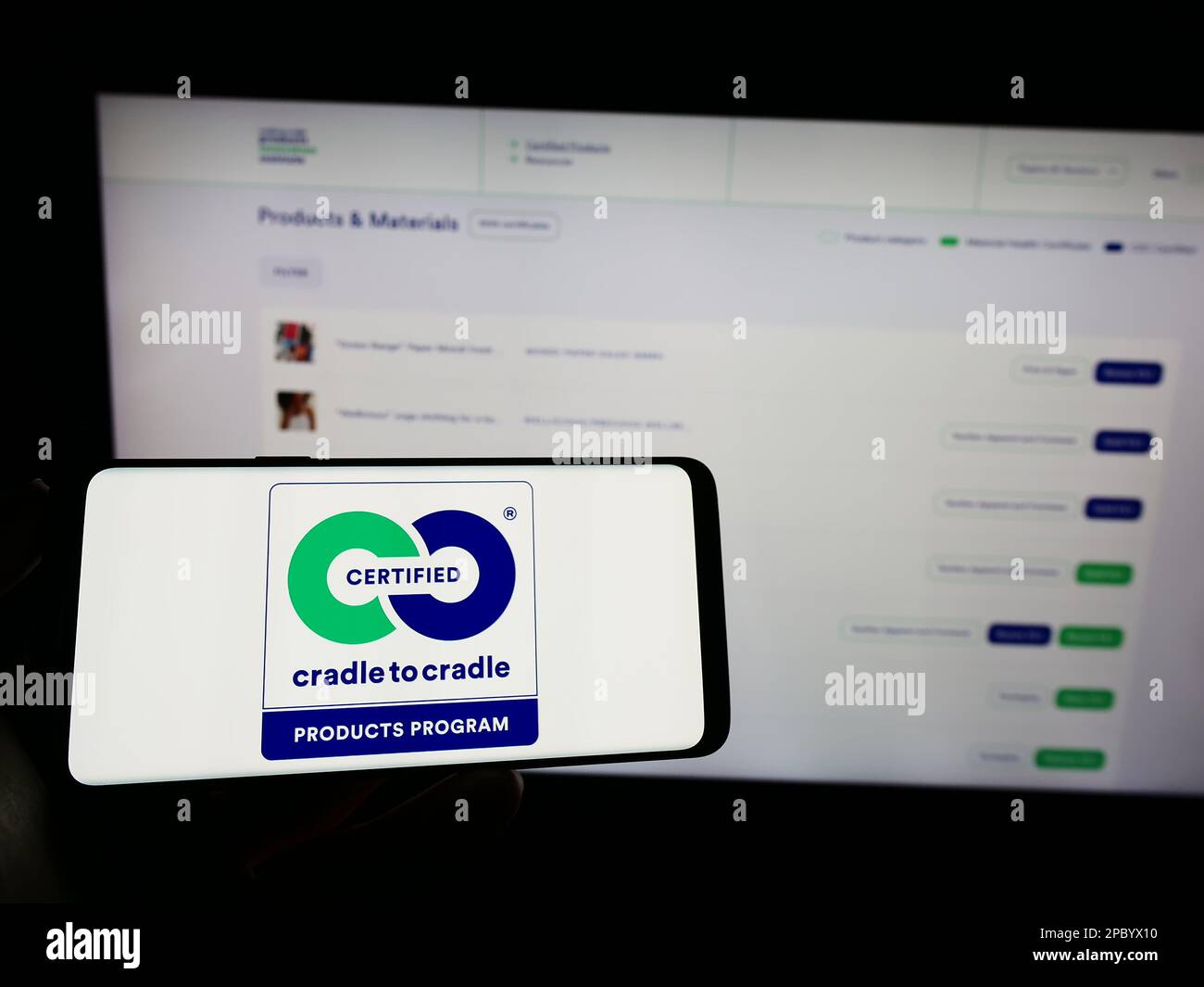 Person holding cellphone with logo of environmental label Cradle to Cradle Certified on screen in front of webpage. Focus on phone display. Stock Photo