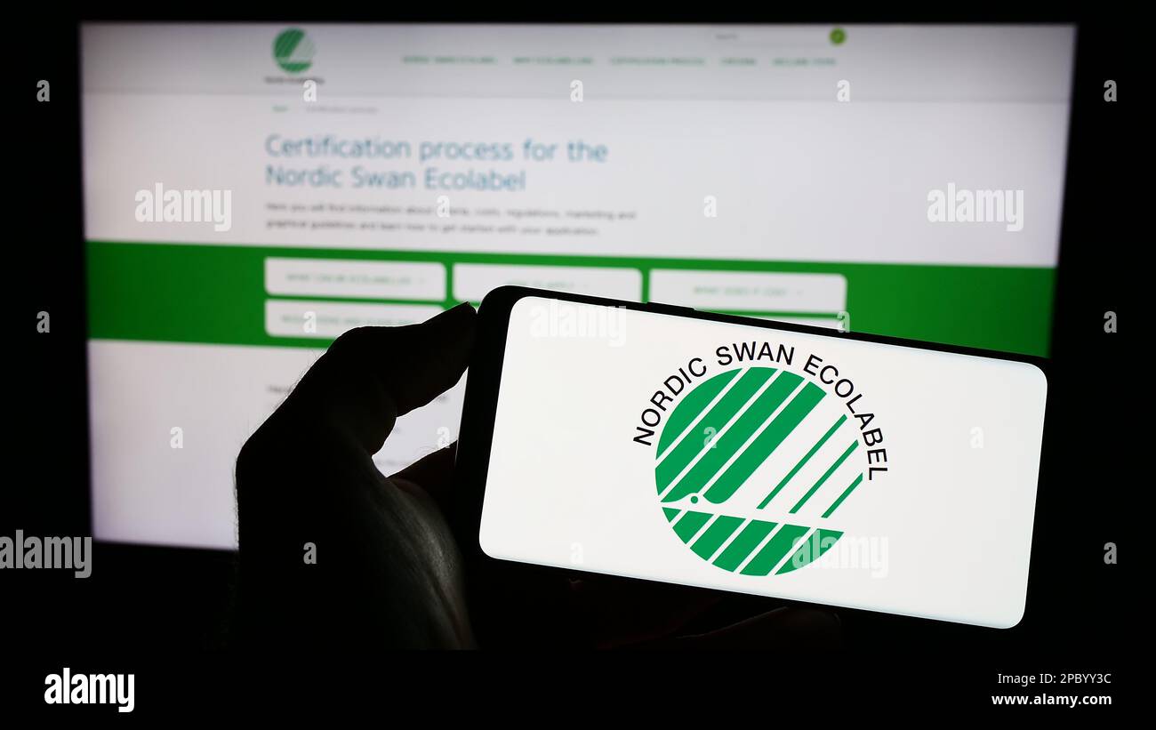 Person holding cellphone with logo of environmental certification Nordic Ecolabel on screen in front of webpage. Focus on phone display. Stock Photo