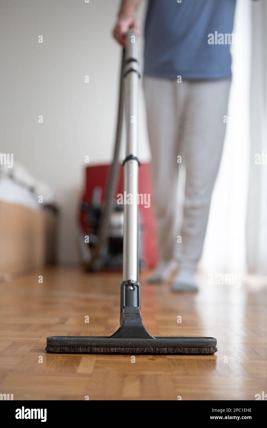 Unrecognizable blurry out of focus male, using a vacuum cleaner brush and extension rod on laminated wooden tile surface inside a generic apartment. C Stock Photo