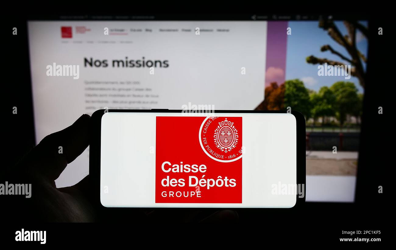 Person holding smartphone with logo of Caisse des Depots et Consignations (CDC) on screen in front of website. Focus on phone display. Stock Photo