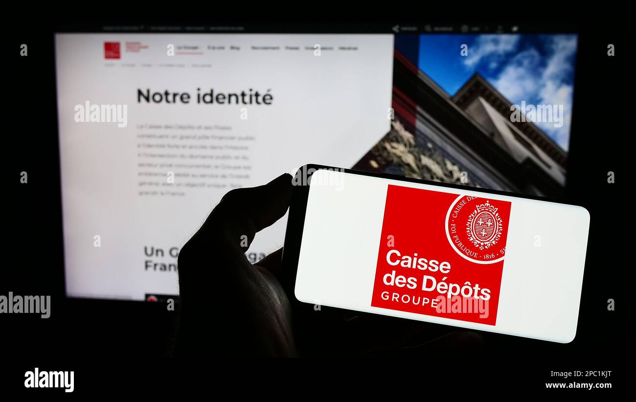 Person holding mobile phone with logo of Caisse des Depots et Consignations (CDC) on screen in front of web page. Focus on phone display. Stock Photo