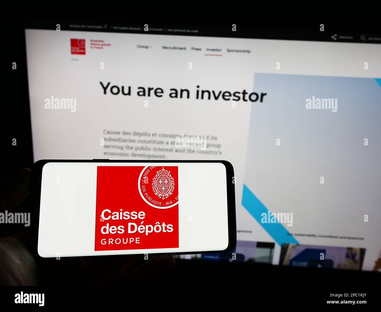 Person holding cellphone with logo of Caisse des Depots et Consignations (CDC) on screen in front of webpage. Focus on phone display. Stock Photo