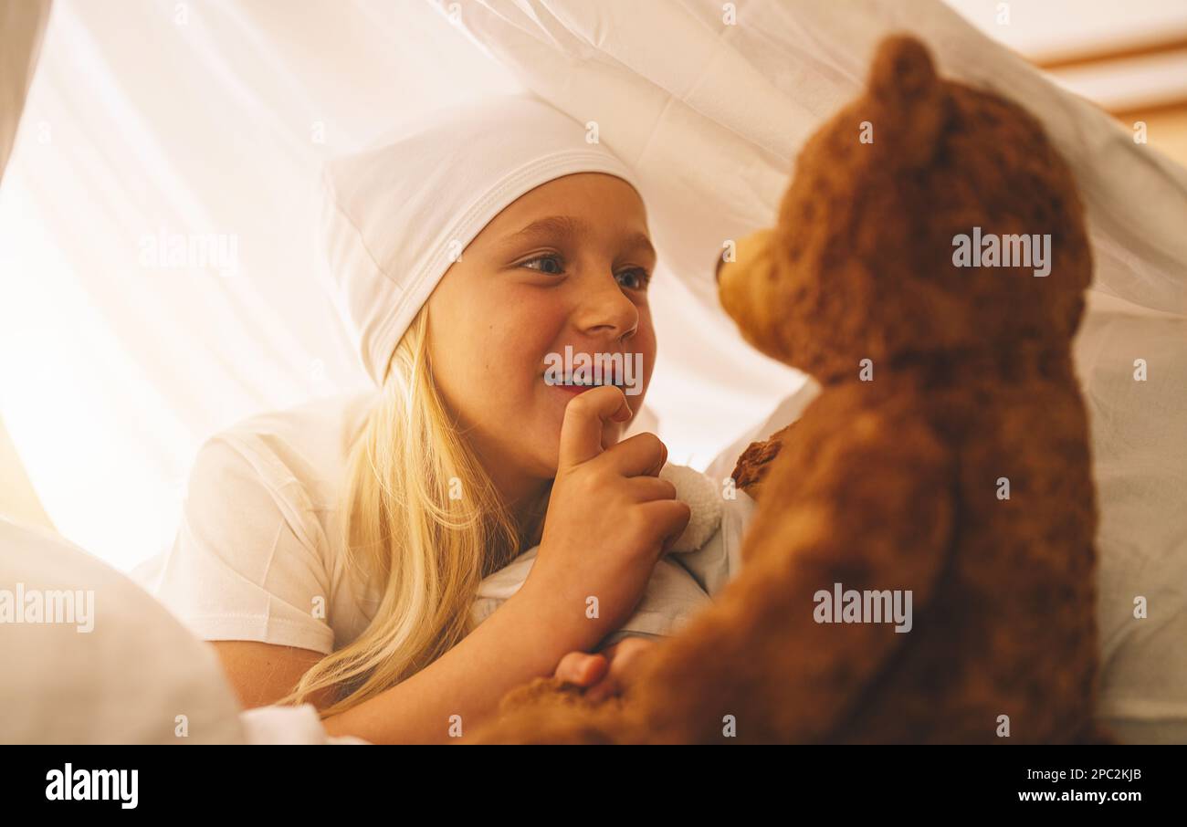 Sweet little girl talking with a teddy bear under blanket at night