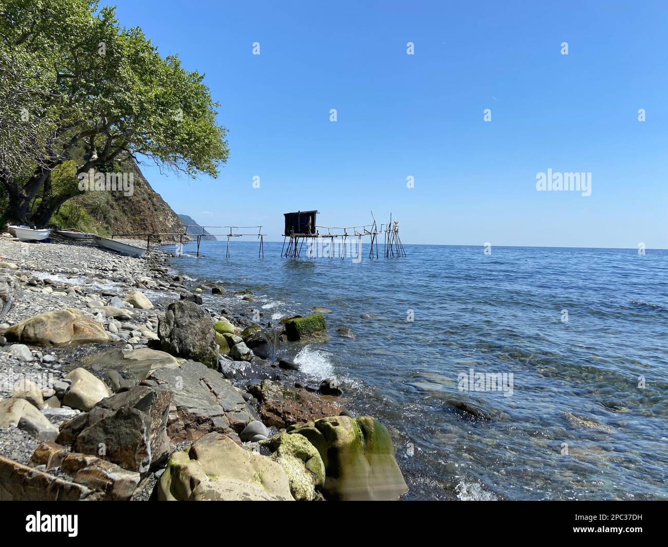 Wooden abandoned stilt tower house fisherman cave on diy pier on the rocky coast shores of fishing village on sunny summer day bright blue skies Stock Photo
