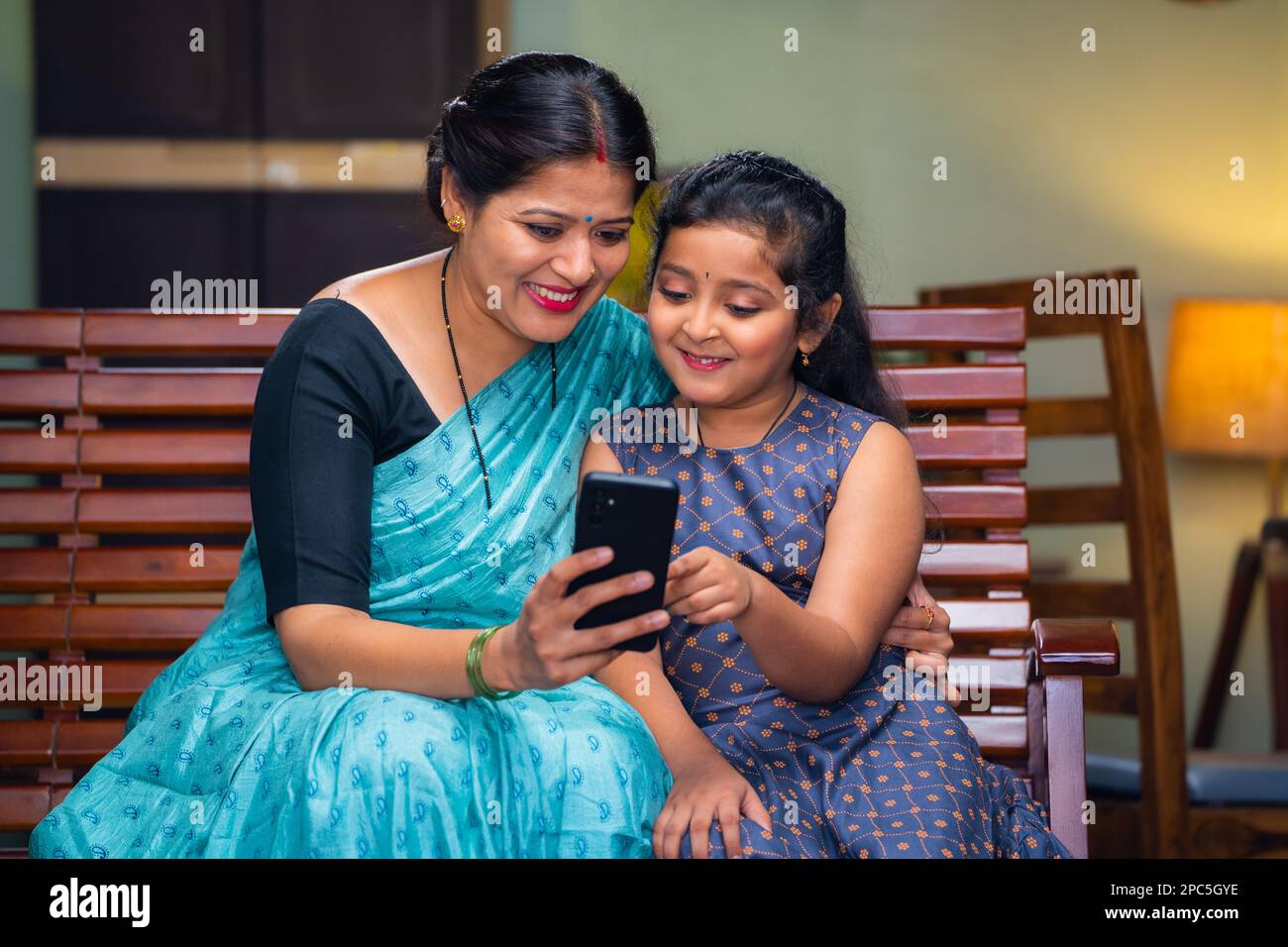 Happy mother showing mobile phone to daughter while sitting on sofa at home - concept of technology, bonding and motherhood. Stock Photo
