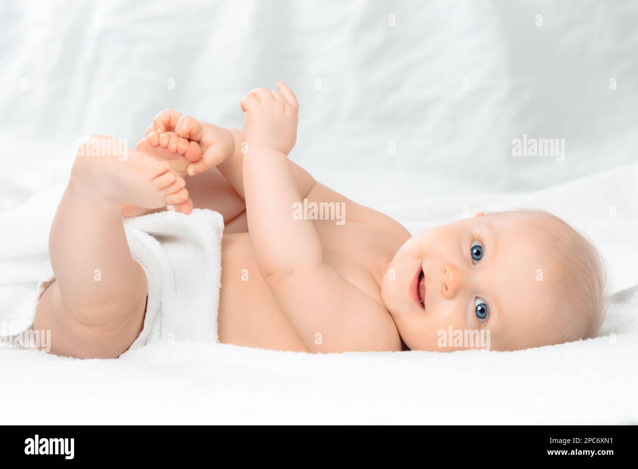 Cute happy baby with blue eyes lying on white bed and holding legs. Little smiling boy looking