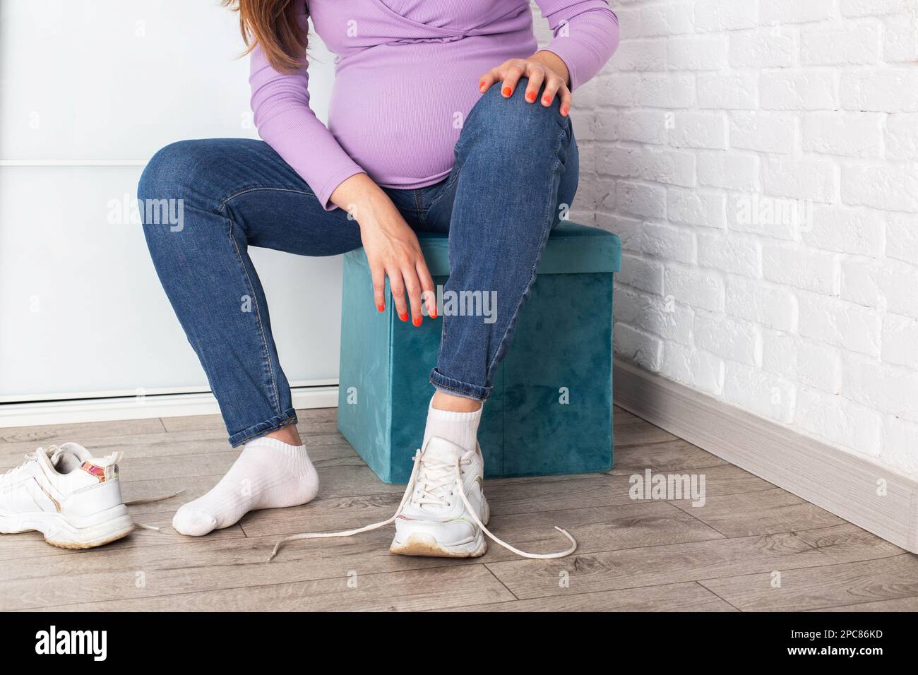 A pregnant girl with a big belly puts on sneakers on her feet. The concept of discomfort and inconvenience when bending over and putting on shoes. Con Stock Photo