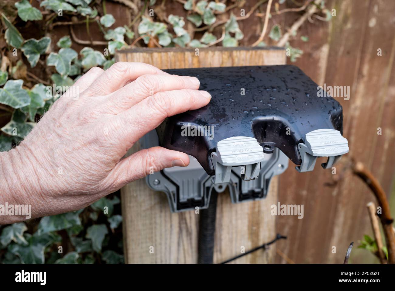 Homeowner about to open the weatherproof cover of a double gang external electrical sockets. Seen in a garden after a heavy downpour Stock Photo