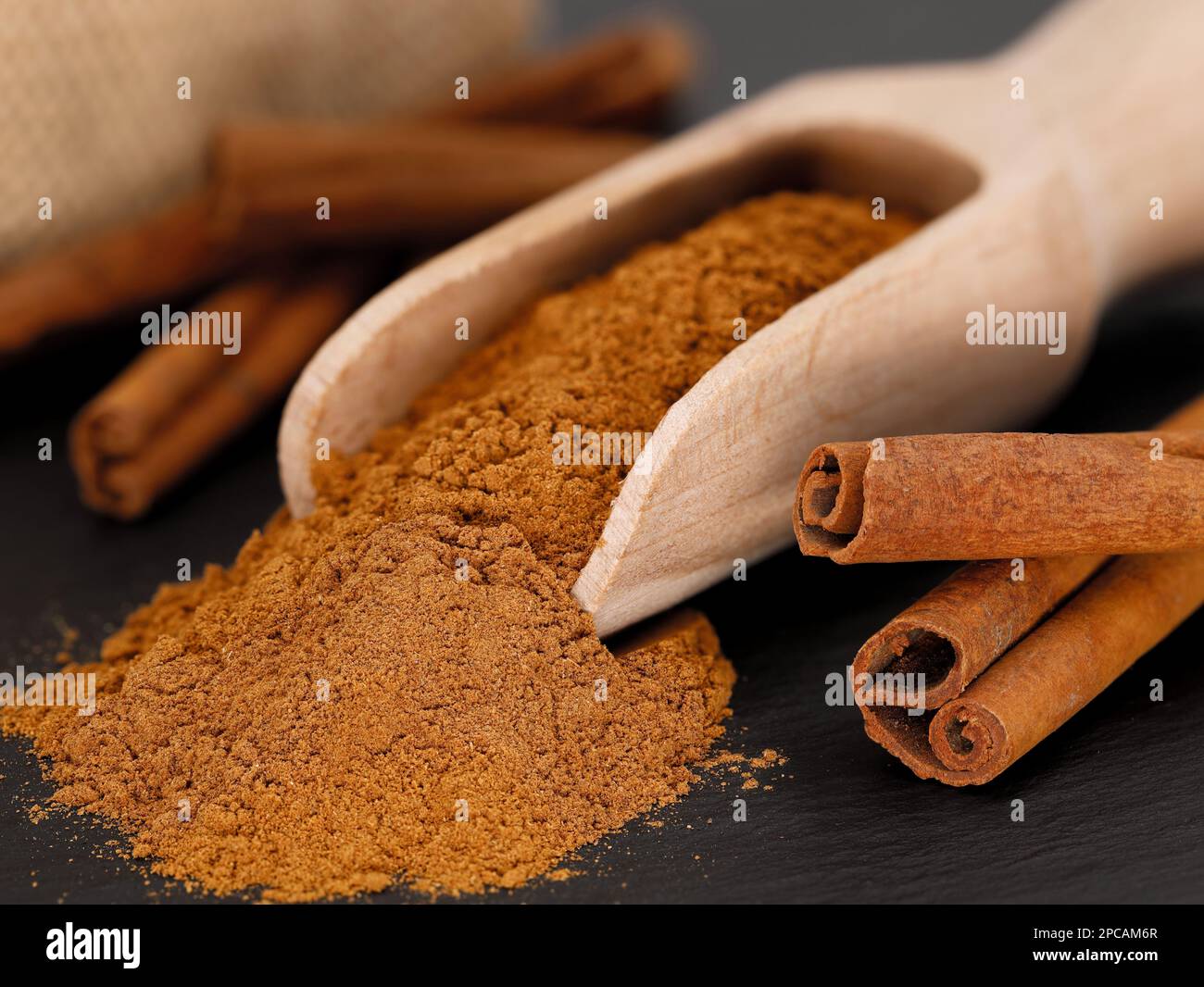 Close up of cinnamon powder in wooden spice scoop next to whole cinnamon sticks on black slate plate Stock Photo