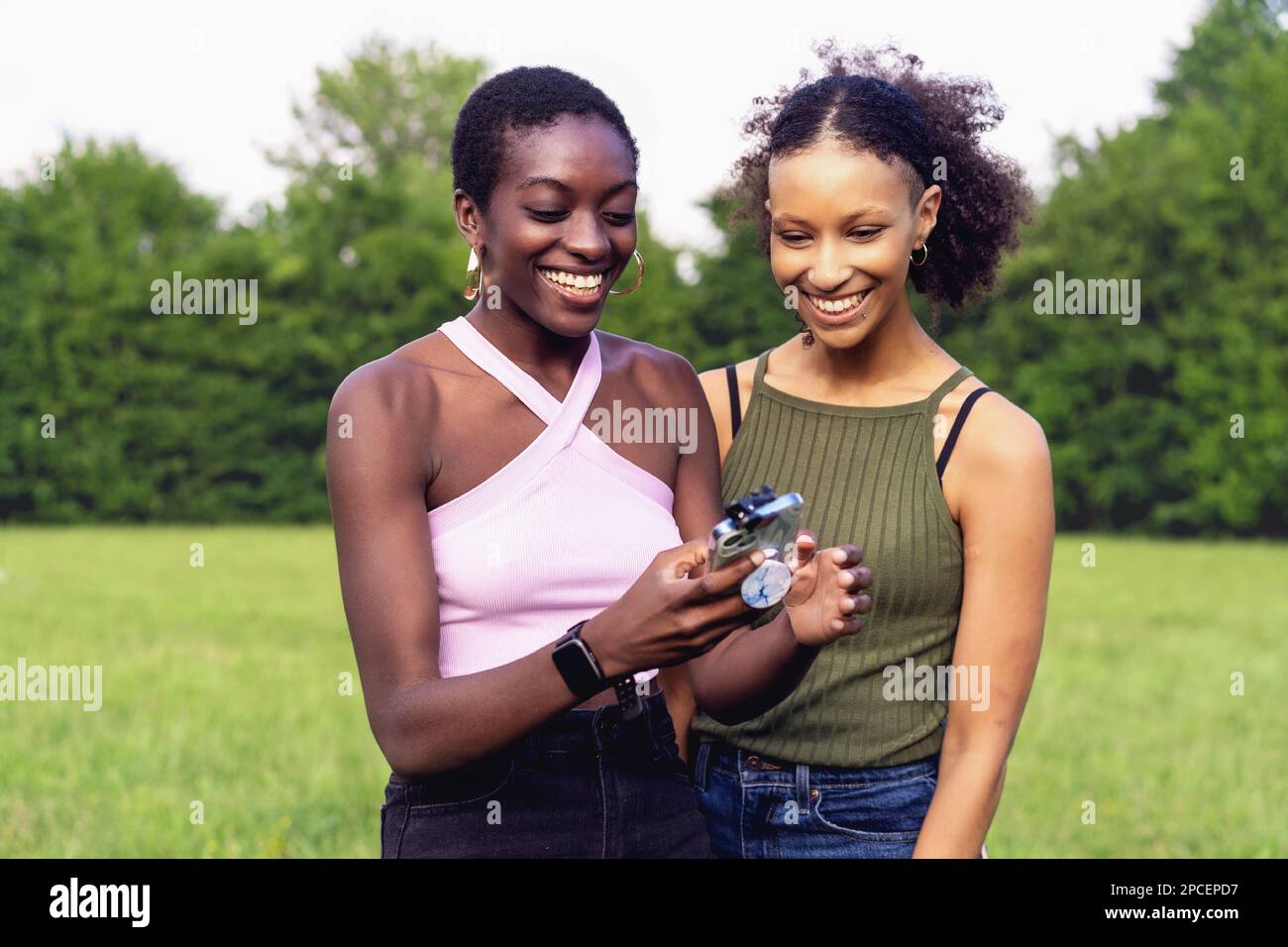 Two young women of color, one with curly long hair and the other with shaved mixed-race hair, enjoy themselves together outdoors in a public park. The Stock Photo