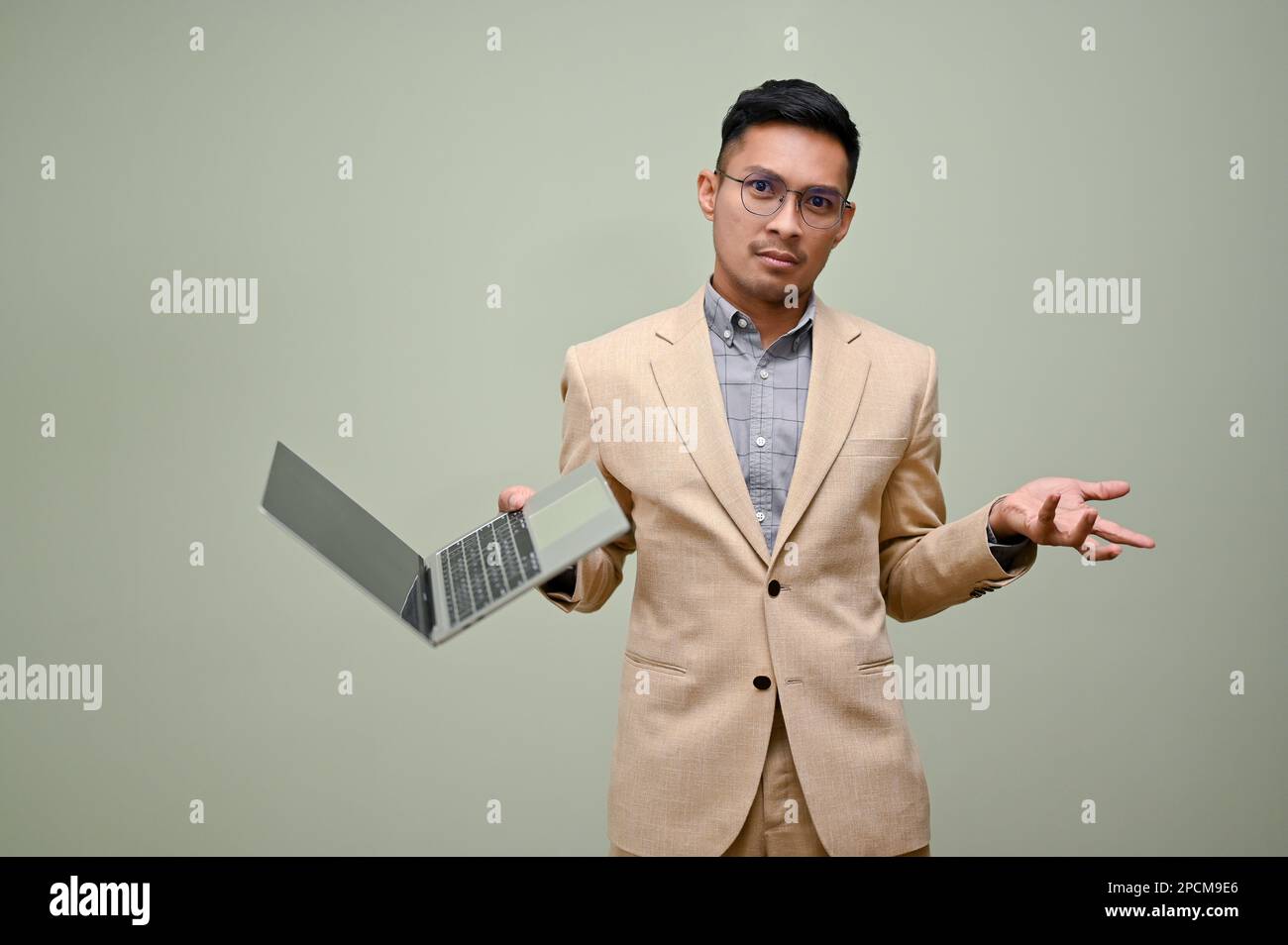 Confused and doubtful millennial Asian businessman in formal business suit stands against a green studio background with his laptop. Stock Photo