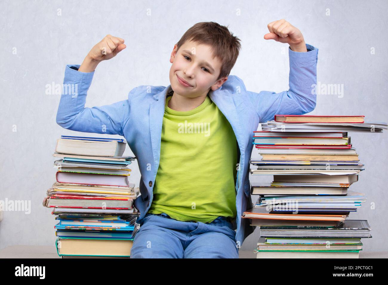 Happy smart school boy sits between stacks of books and shows a sign of strength.