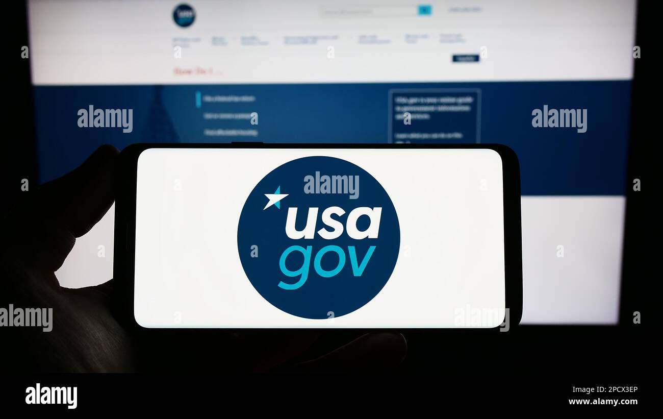 Person holding smartphone with logo of US federal government website USA.gov on screen in front of website. Focus on phone display. Stock Photo