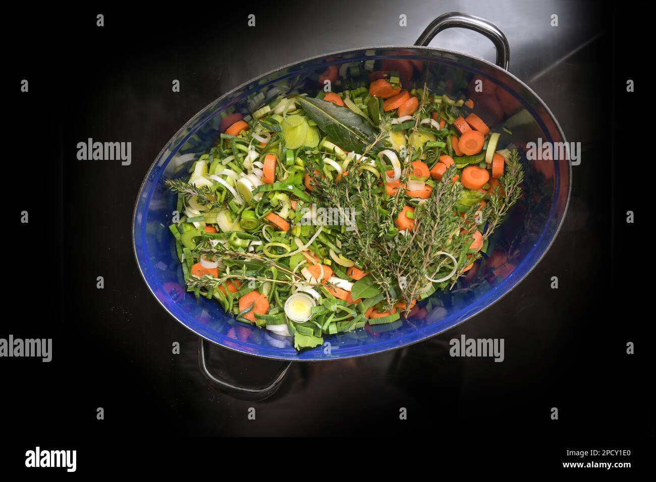 Chopped raw vegetables like carrots, celery and leek with herbs like thyme and bay leaves in a dark blue roaster on the black induction stove top, for Stock Photo