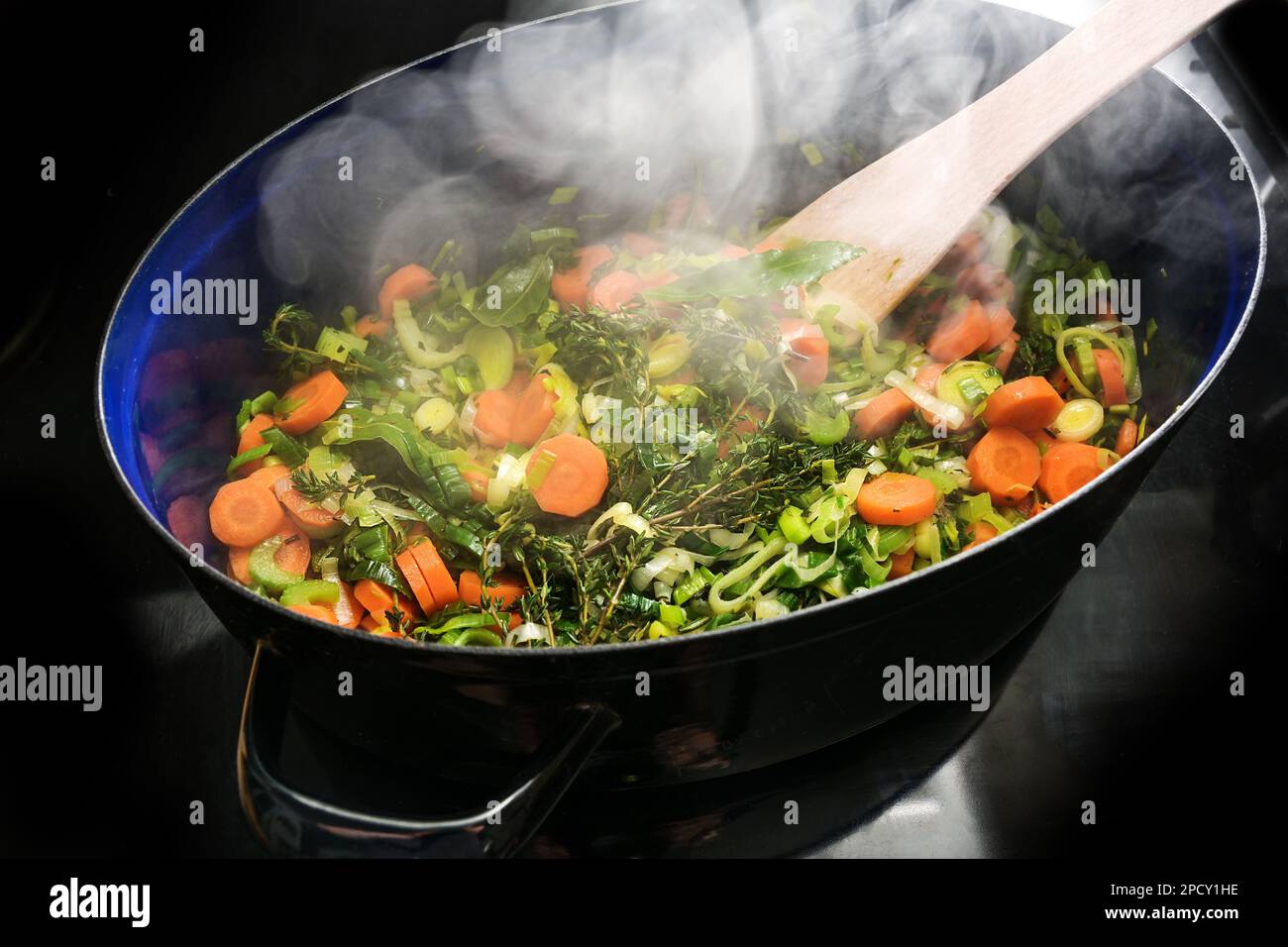 Steaming vegetables and herbs in a cooking pan on the black stove top, ingredients for a soup like carrots, celery, leek, thyme and bay leaves, copy s Stock Photo