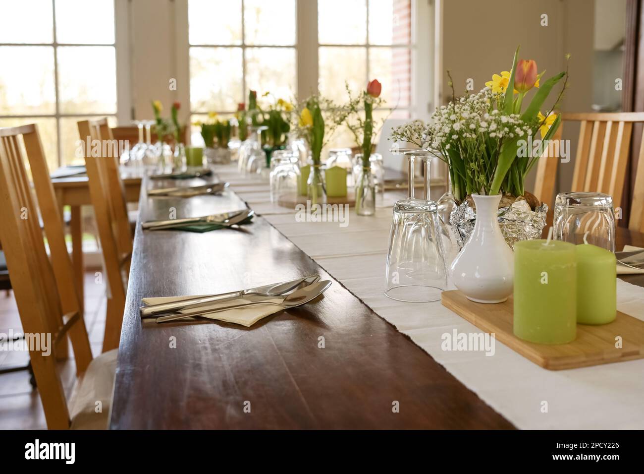 Large wooden table decorated for a dinner party with spring flowers, green candles and various drinking glasses on a light table runner in a dining ro Stock Photo