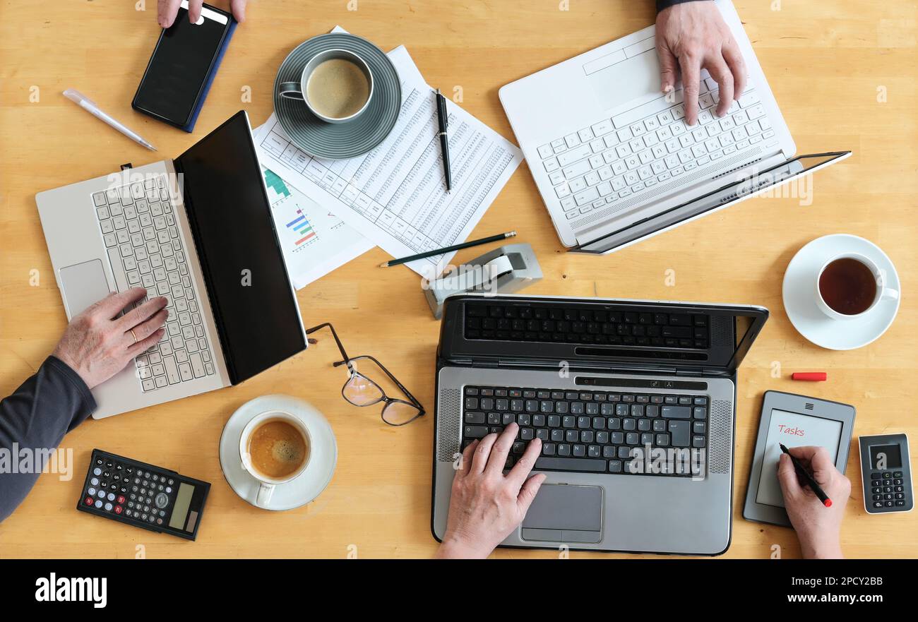 Working together on a joint business project, top view of the wooden table with hands, three laptops, papers, calculator and coffee, high angle shot f Stock Photo