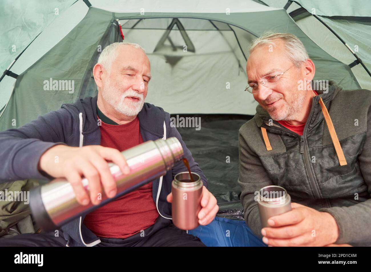 Elderly man pouring tea by male friend in camping tent during vacation at forest Stock Photo
